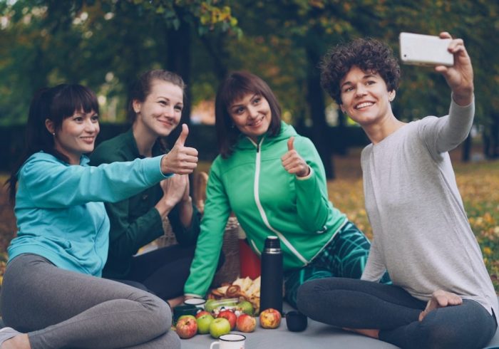 Attractive young ladies yogini are taking selfie using smartphone during picnic in park in autumn. Girls are posing and smiling having fun sitting together on mats.